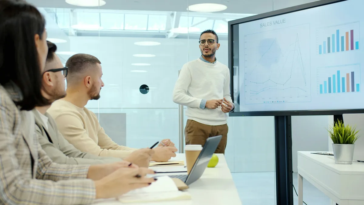 A presenter beside a screen of sales charts speaking to colleagues seated around a meeting table