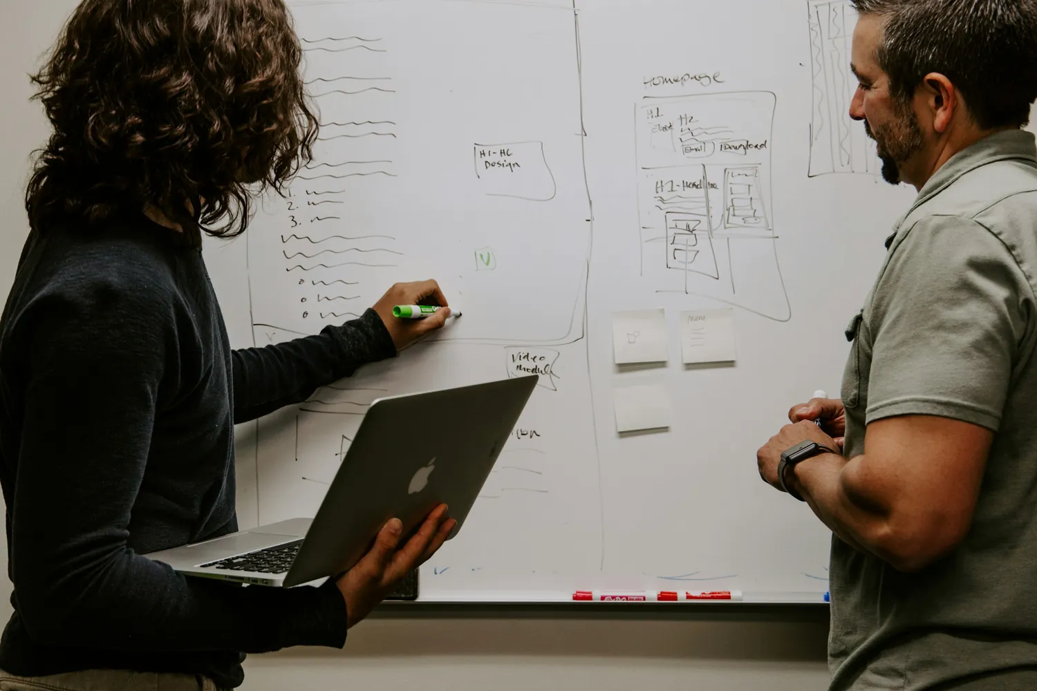 Two colleagues at a whiteboard covered in page-layout sketches, one writing while holding a laptop