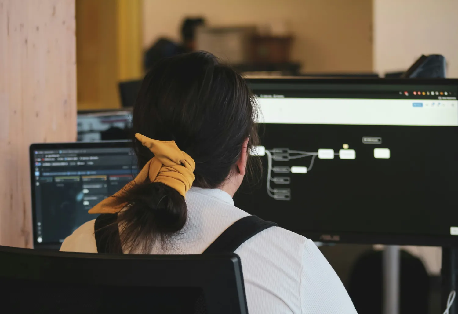 A developer seen from behind at a laptop and a widescreen monitor showing a node-based workflow diagram