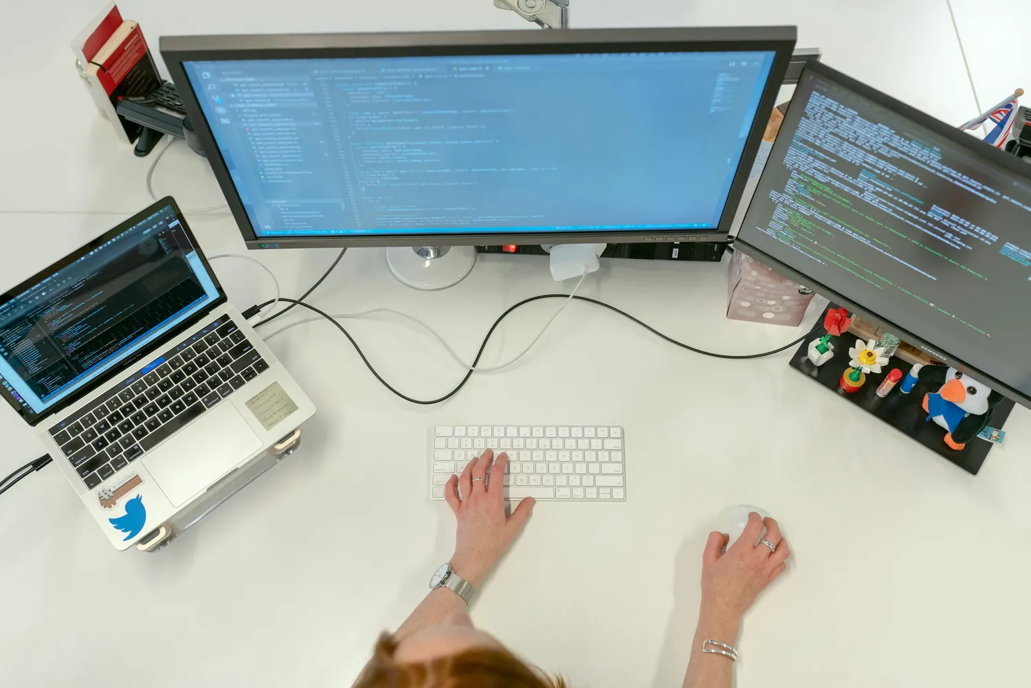 Overhead view of a developer at a white desk, hands on keyboard and mouse between a laptop and two monitors of code