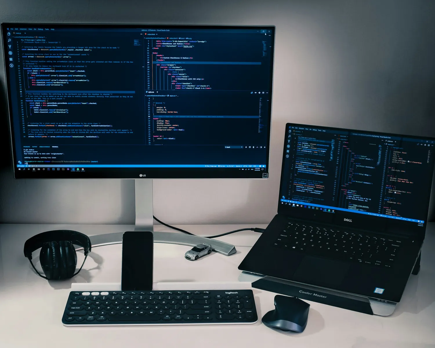An ultrawide monitor and a laptop side by side, both showing HTML, CSS and JavaScript in a code editor