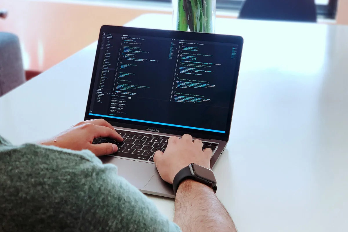 Developer typing on a MacBook Pro showing a code editor at a bright white desk