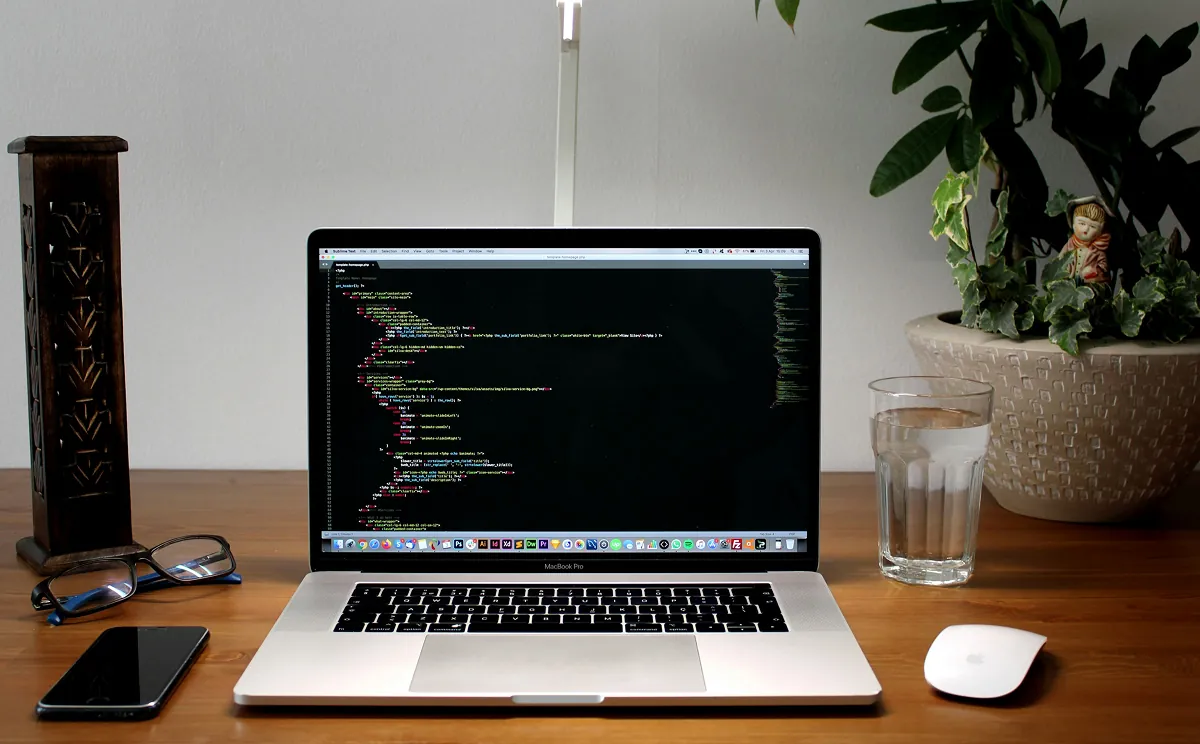 MacBook Pro showing code on a wooden desk beside glasses, a phone and a potted plant