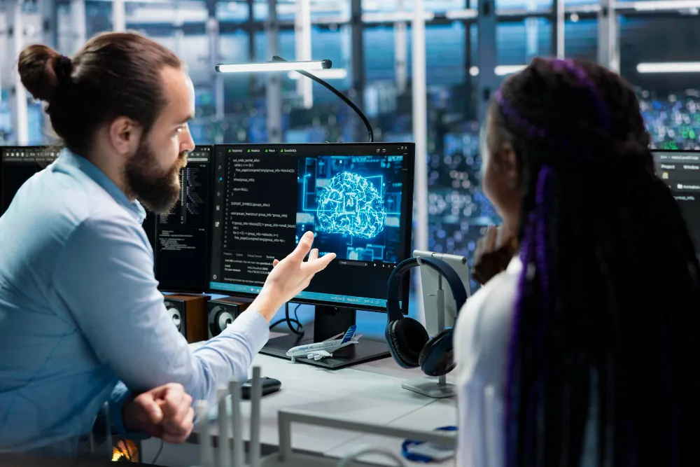 Two engineers discussing a glowing AI brain visualisation on a monitor in a server room