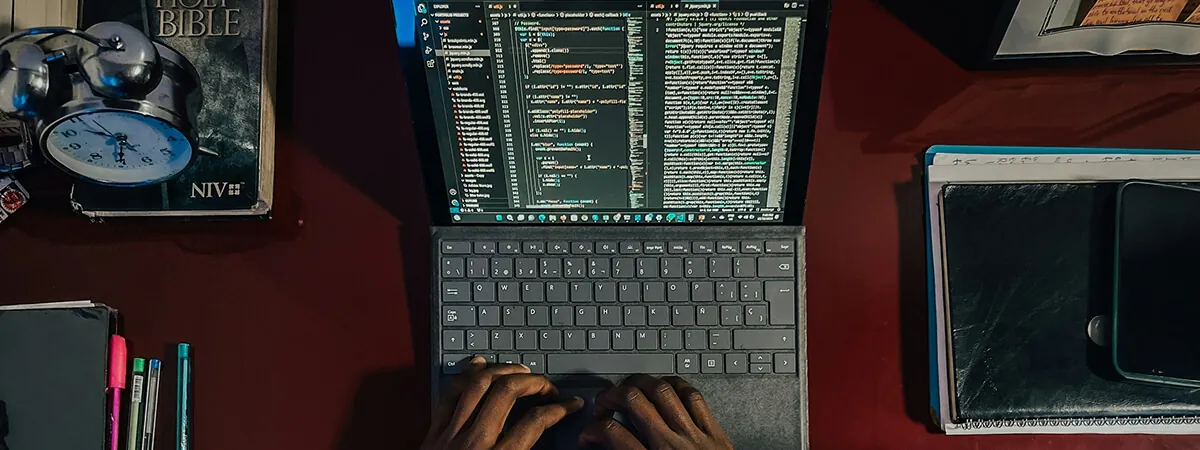 Overhead view of hands typing on a laptop showing a code editor, beside a notebook and alarm clock