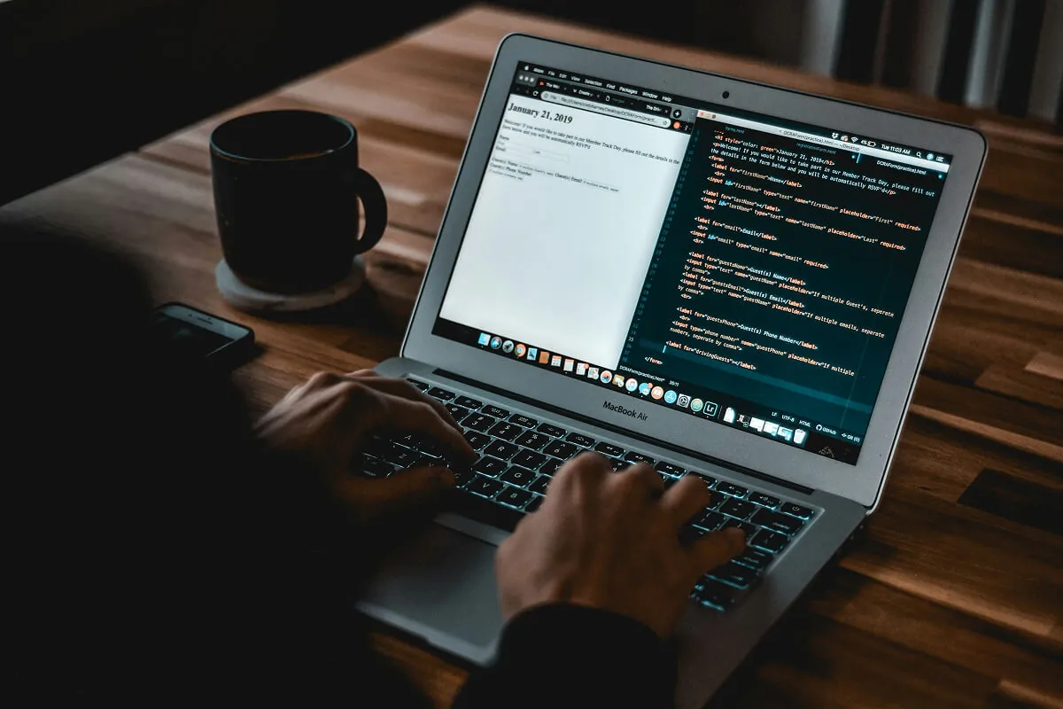 Developer typing on a laptop showing a code editor beside a coffee mug on a wooden desk