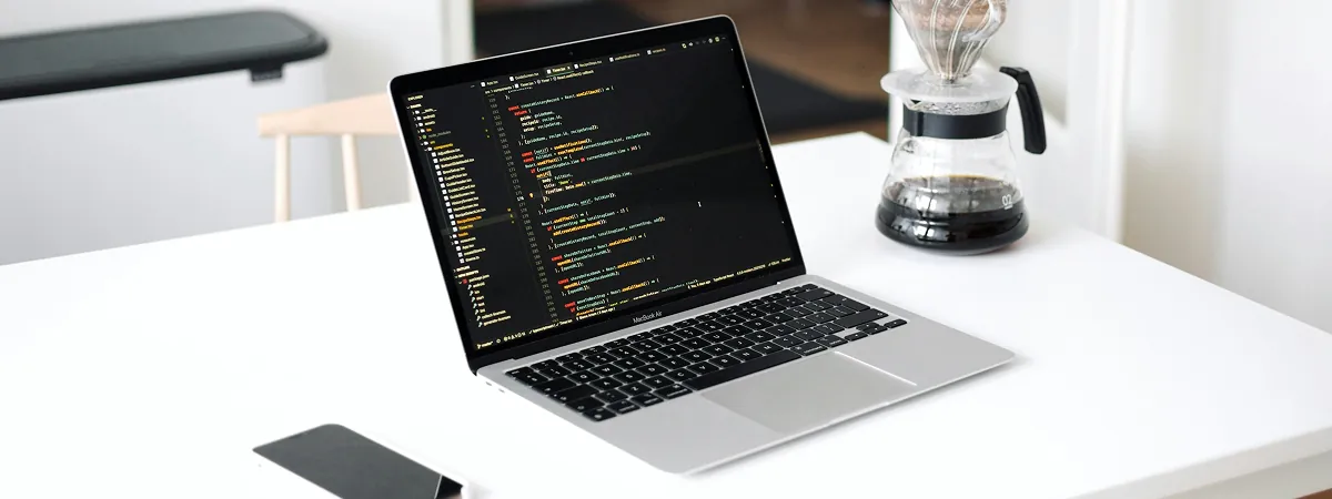 Laptop displaying code on a bright white desk beside a pour-over coffee carafe