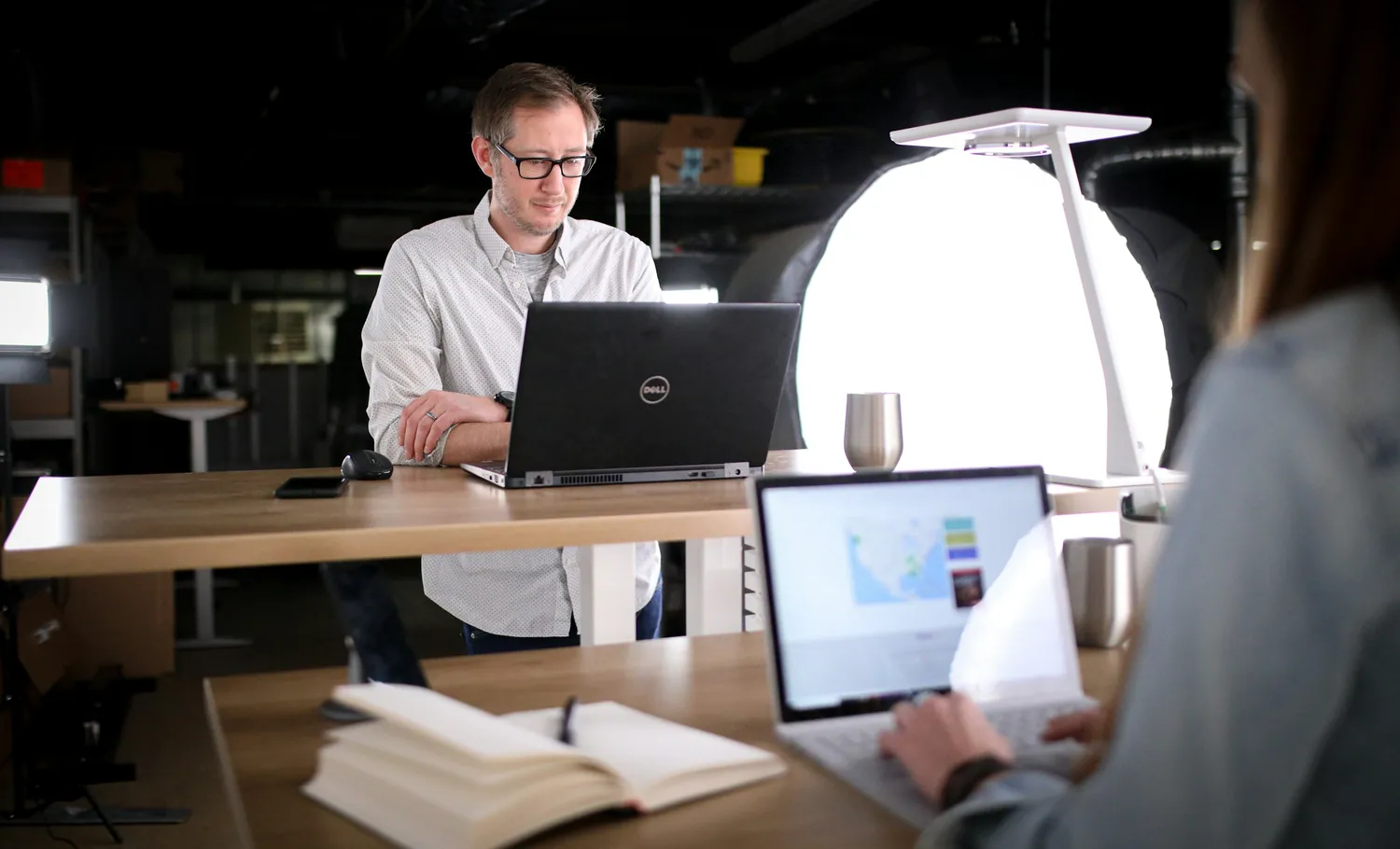 Two colleagues working on laptops at standing desks in an open-plan studio