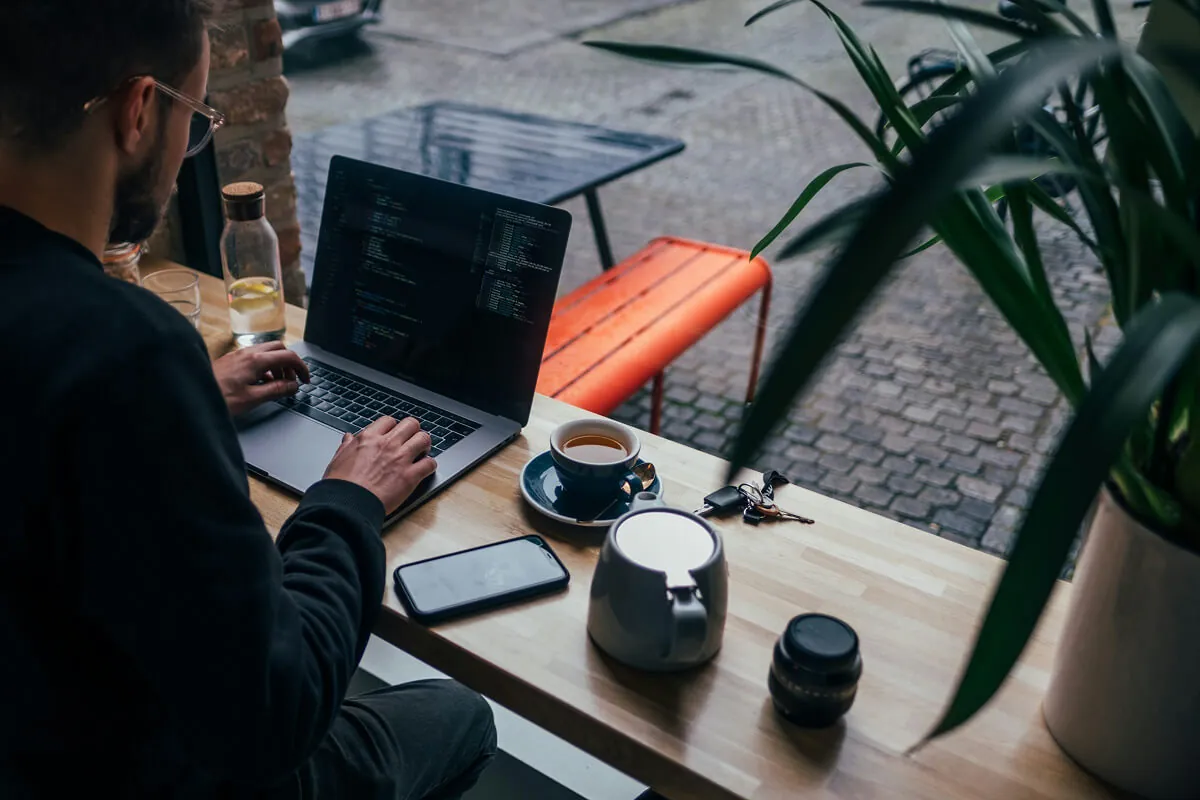 Developer coding on a laptop at a cafe table with a coffee cup, phone and plant nearby