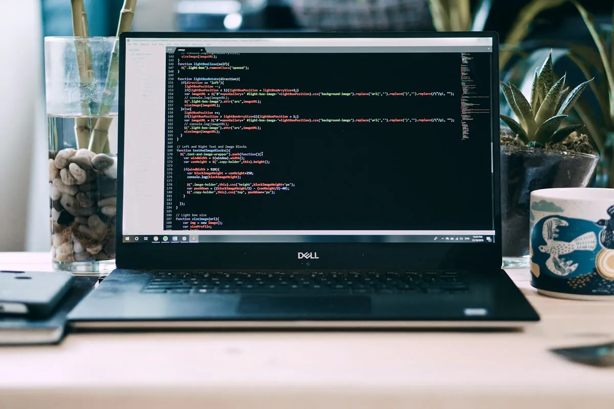 Dell laptop on a desk showing JavaScript source in an editor, beside a plant and a mug