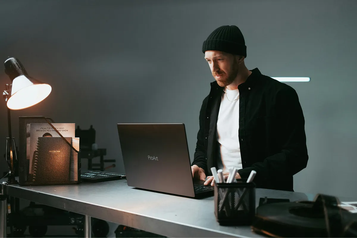 Developer working at a laptop on a metal workbench under a desk lamp in a dimly lit studio