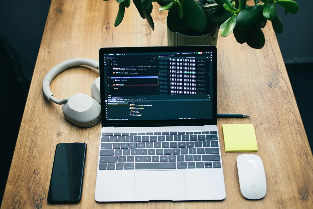 Laptop open on a wooden desk showing a code editor, with headphones, phone, mouse and a plant