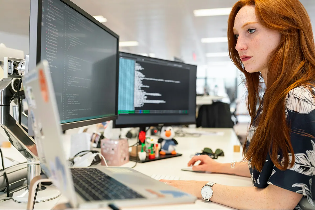 A developer in an open-plan office working across a laptop and two monitors of code