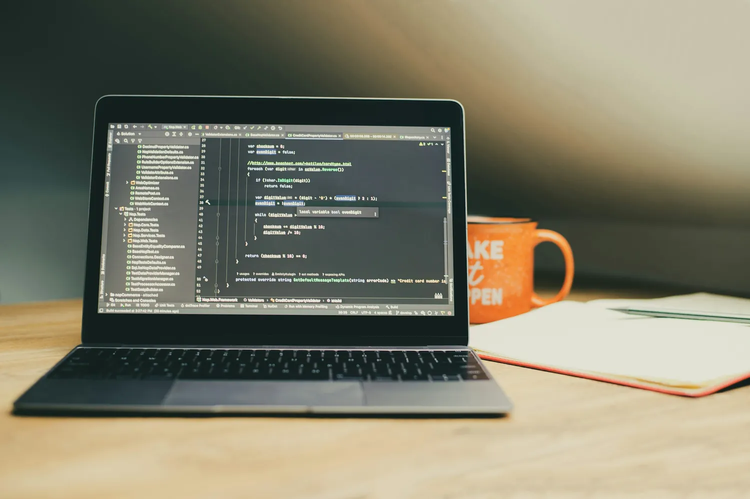 A laptop on a wooden desk showing C# validation code in an IDE, beside a notebook and an orange mug